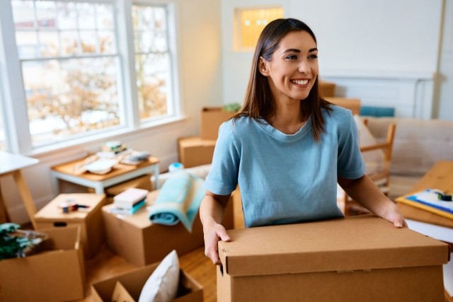 A smiling young woman carries moving boxes into her new apartment.