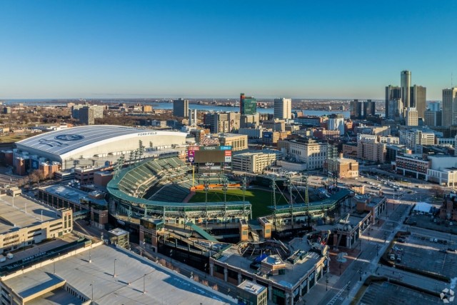 An aerial view of Detroit shows the downtown skyline and Comerica Park in the foreground and the Detroit River and Ontario, Canada, in the background.