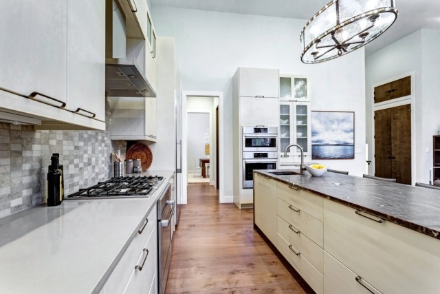 A view of a kitchen in a modern apartment shows granite countertops, stainless steel appliances, tile backsplash, and a unique light fixture.