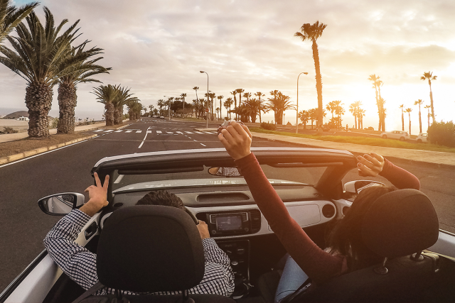 A couple on a road trip at sunset - the car is a convertible model that is driving down a road lined with palm trees