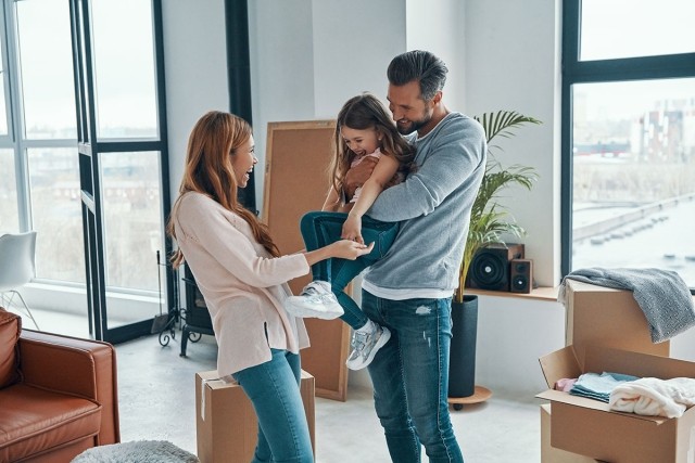 Man holding kid up with woman holding the kid's hands as they all laugh in new apartment.