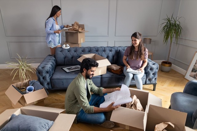 Two women and a man who are roommates unpacking boxes in their new apartment living room.