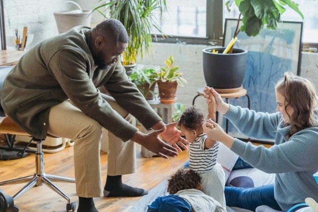 Parents play with their two young children on the floor of a sunlit apartment.