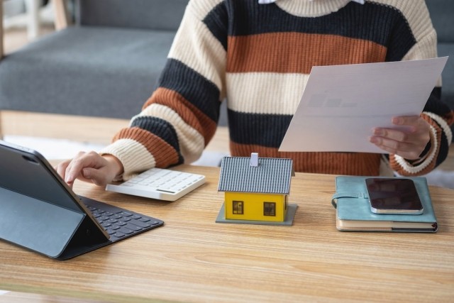 Woman typing on laptop while holding papers with a calculator, model house, phone, and notebook in front of her.